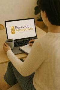 A person sitting at a wooden table with a mug, notebook, and open laptop displaying the Renewed Perspectives Therapies & Life Coaching logo.